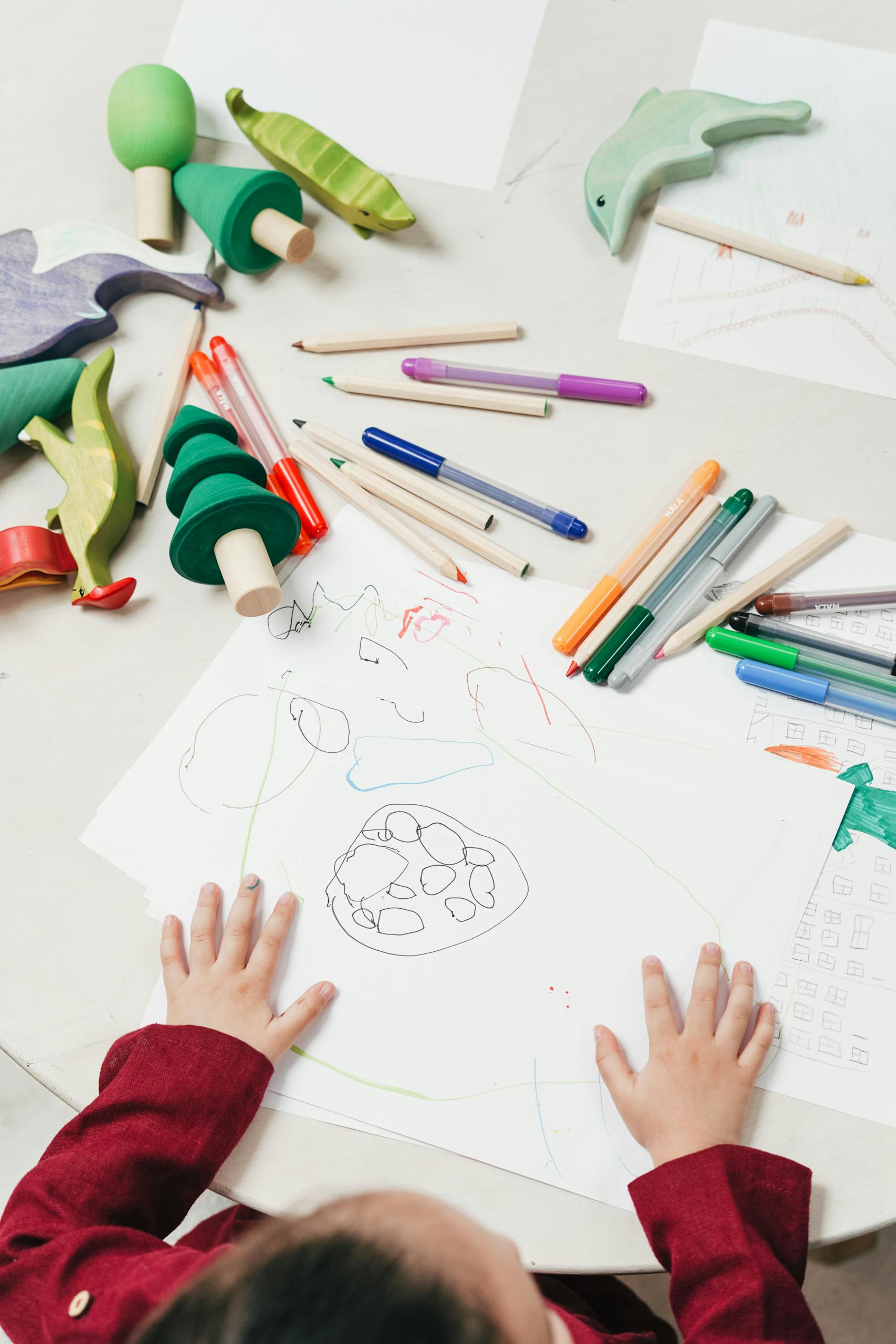 A child engages in creative drawing with colored pencils surrounded by wooden toys on a table as they homeschool in kindergarten.