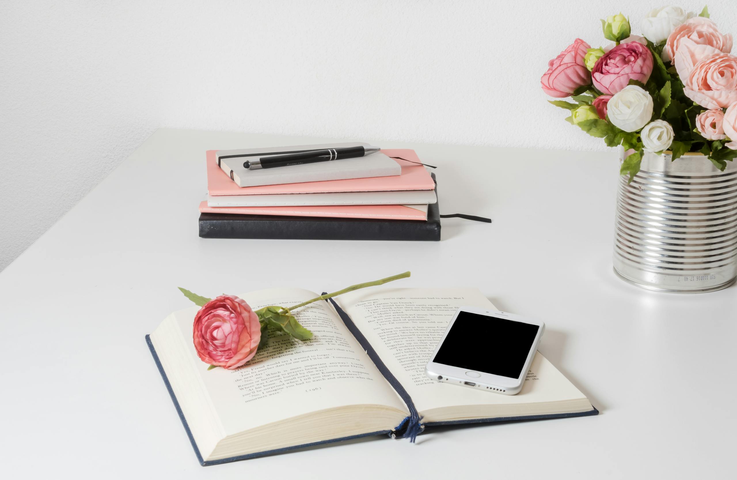 A stylish home office setup with open book, smartphone, and flowers on a white desk with a stack of homeschool books in the background