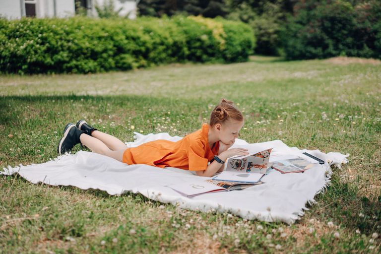 Young girl reading a book on picnic blanket in a sunny park. Homeschooling over the summer