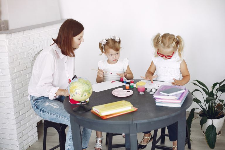 Small kids attentively painting in kindergarten with adult assistance while sitting together at round table doing homeschool lessons